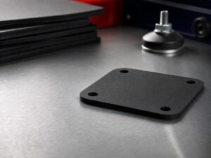 Matte black precision-cut neoprene pad on an industrial workbench with rubber sheets and an isolation mount in the background.